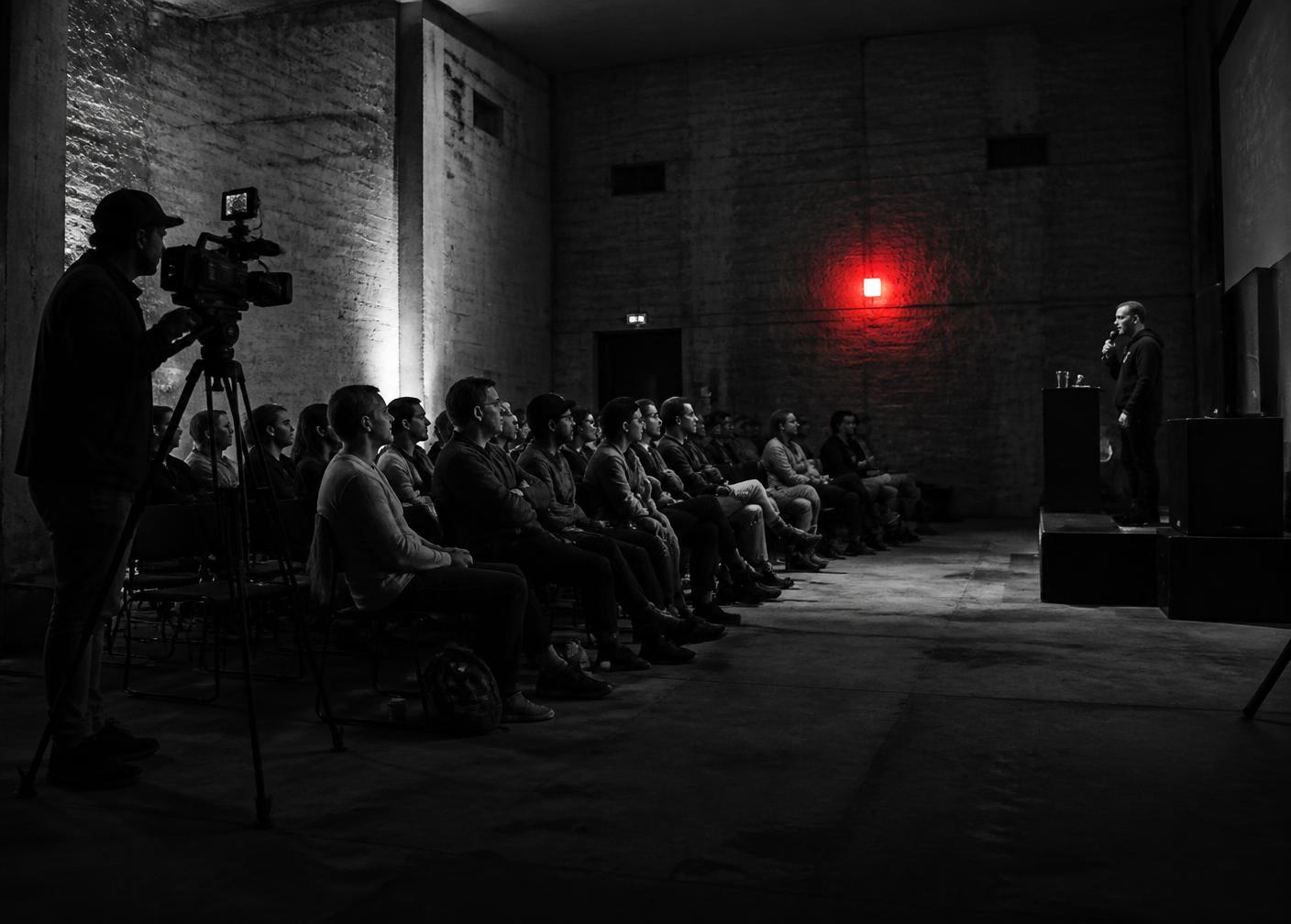 An audience seated in a dark concrete room listening to a speaker while a camera operator films