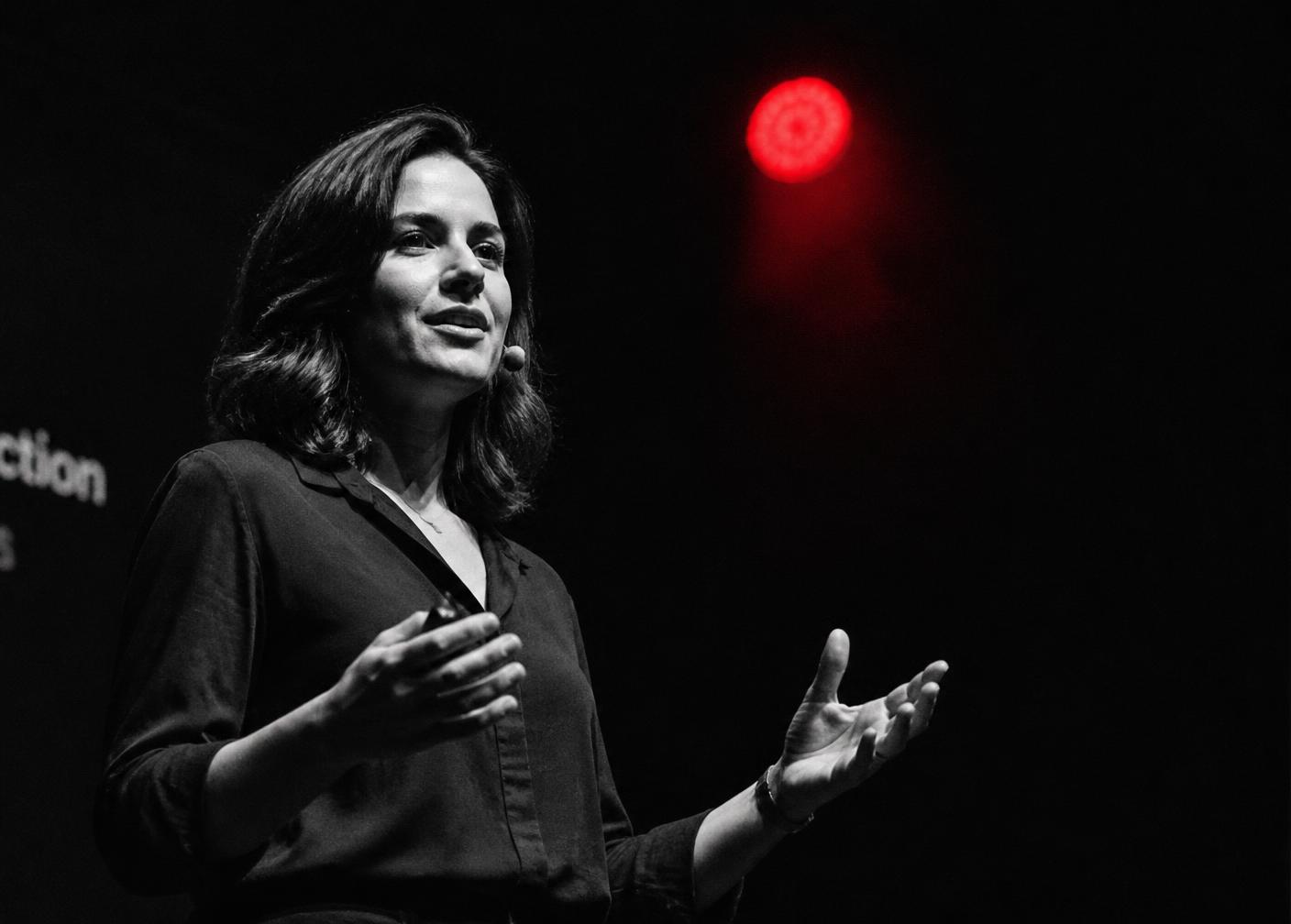 A woman speaking on a dark stage under a single spotlight with a red stage light behind